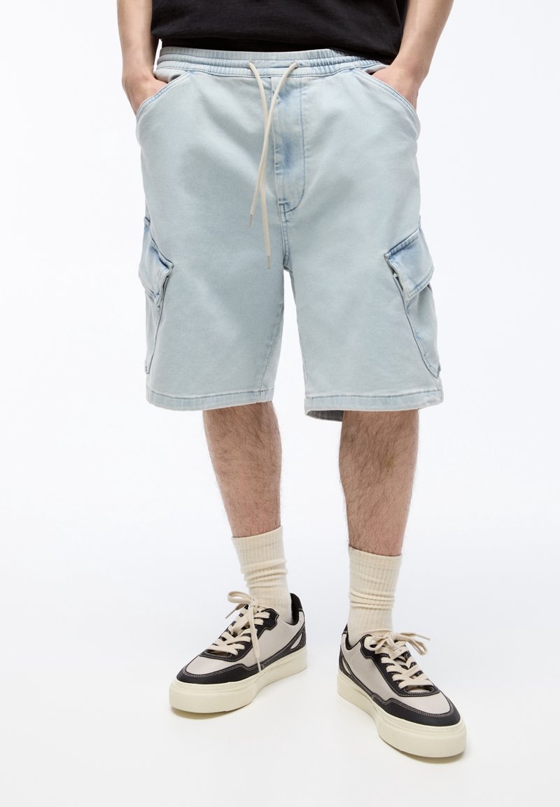 Short en denim bleu clair avec une taille à cordon, deux poches latérales et des poches cargo sur chaque jambe, associé à des baskets noires et beige.