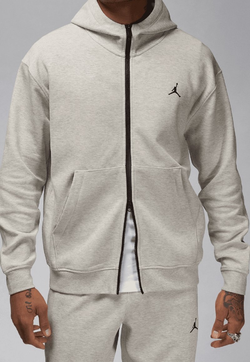 Homme portant un sweat à capuche zippé gris clair et un pantalon de survêtement assorti avec le logo Jumpman noir sur la poitrine et la jambe, debout devant un fond gris.