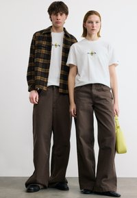 Δύο μοντέλα φορούν λευκά μπλουζάκια με την επιγραφή «New York City» και φαρδιά καφέ παντελόνια, το ένα με ένα καφέ καρό σακάκι και το άλλο κρατώντας μια κίτρινη τσάντα.