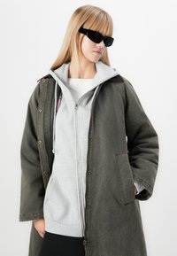 Lungo cappotto grigio scuro con hardware dorato sopra una felpa grigia con zip. Occhiali da sole neri. Tessuto liscio, vestibilità comoda, senza fantasie.
