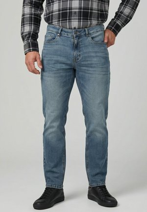 Uomo con jeans azzurri, scarpe da ginnastica nere e camicia a quadri nera e grigia a maniche lunghe, in piedi con una mano in tasca.
