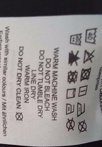 Étiquette avec les instructions d'entretien : tissu blanc, texte noir. Symboles pour lavage en machine, pas de blanchiment, sécher à plat, repasser à température tiède, ne pas laver à sec.