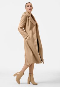 Beige wollen blend lange jas met zijzakken, notch-kraag en een open voorkant. Model draagt beige krokodillenhuid-embossed enkellaarsjes met een blokhak.