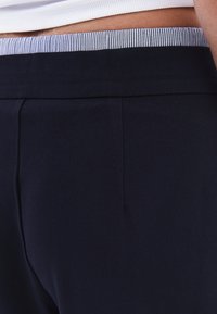 Pantaloni blu navy con una texture liscia, caratterizzati da una vita a righe in contrasto e un design su misura con due pieghe frontali.