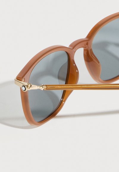 Καφέ γυαλιά ηλίου με στρογγυλούς γκρι φακούς και χρυσά μεταλλικά πλαίσια, εμφανιζόμενα σε κοντινό πλάνο πάνω σε λευκή επιφάνεια.