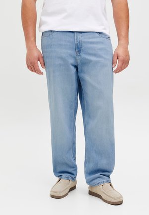 Homme portant un jean ample bleu clair, des chaussures beiges à enfiler et un t-shirt blanc, debout devant un fond clair uni.