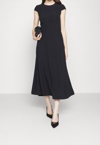 Robe noire à manches courtes avec un corsage ajusté et une jupe évasée. Le modèle tient une pochette noire et porte des escarpins noirs avec un accent doré.