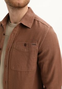 Rjava flanelasta srajca z teksturirano površino, s sploščeno žepom in koničastim ovratnikom. Nosi se preko svetlobežnega zgornjega dela.