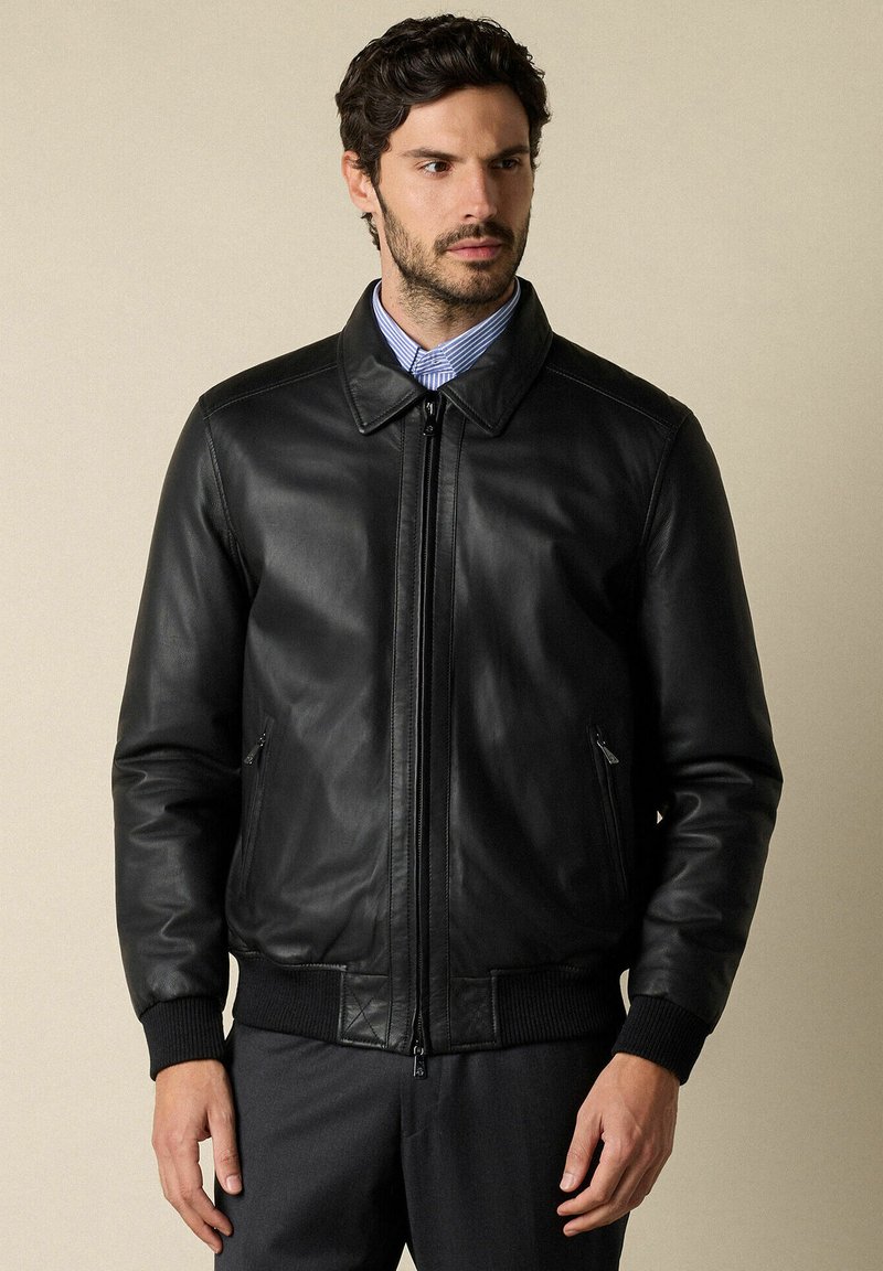 Man in een zwart leren ritsjack over een blauw gestreept overhemd met kraag, staand tegen een effen beige achtergrond.