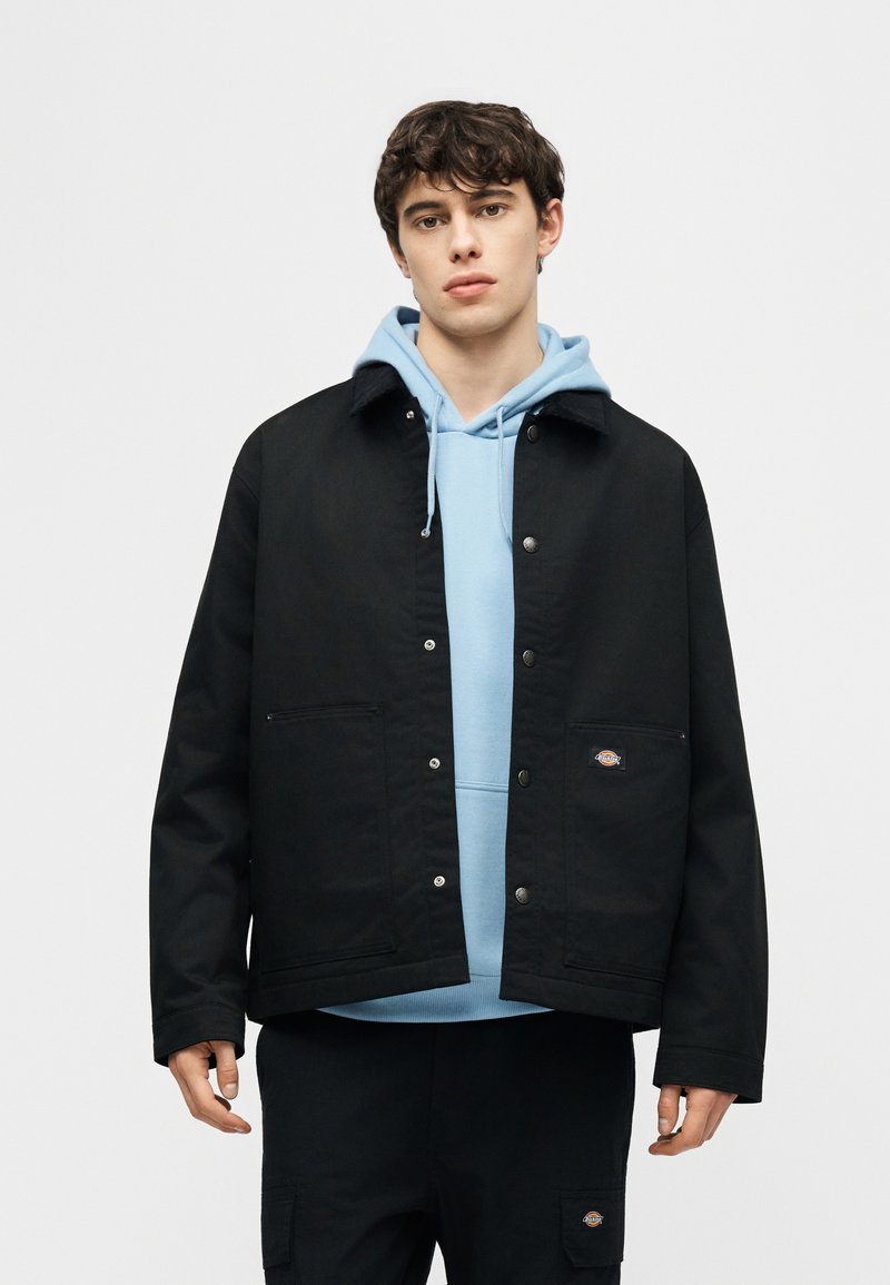 Giovane uomo indossa una felpa con cappuccio azzurra sotto una giacca nera con bottoni e toppe con logo su uno sfondo chiaro e semplice.