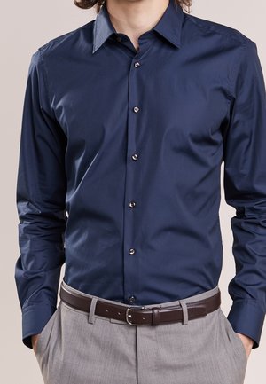 Mann trägt ein eng anliegendes dunkelblaues Hemd mit Knöpfen, eingesteckt in hellgraue Hose mit braunem Ledergürtel, Hände in den Taschen.