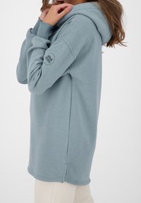 Vaaleansininen ylisuuri huppari, jossa on kengurupocket, ribbed-hihansuut ja logotyyppi hihassa. Puuvillasekoitekangas, jossa on sileä pinta.