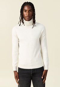 Suéter blanco de cuello alto en una suave tela de punto, diseño ajustado, mangas largas, puños y dobladillo acanalados, combinado con jeans oscuros texturizados.