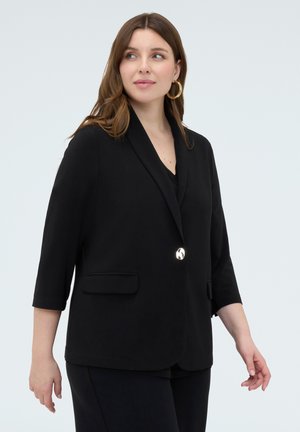 Frau trägt schwarzen Blazer mit einem silbernen Knopf, Dreiviertelärmeln und goldenen Creolen, blickt zur Seite.