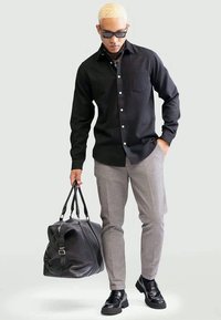 Chemise noire à boutons, pantalons gris à carreaux, chaussures noires et un grand sac de sport noir. Tissu lisse, coupe standard et design structuré.