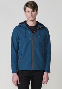 Giacca softshell blu con cappuccio, dotata di tasche con zip, cordini regolabili e una consistenza morbida. Dettaglio della zip a contrasto.