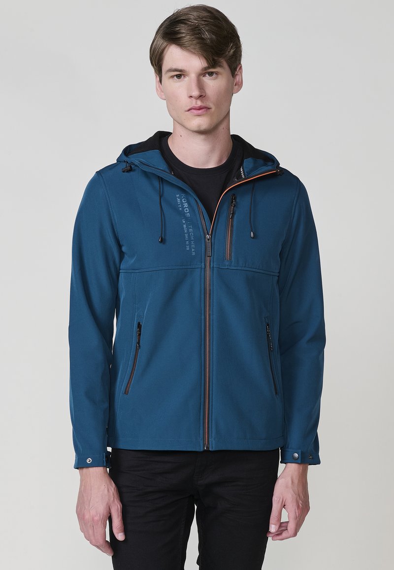 Giacca softshell blu con cappuccio, dotata di tasche con zip, cordini regolabili e una consistenza morbida. Dettaglio della zip a contrasto.