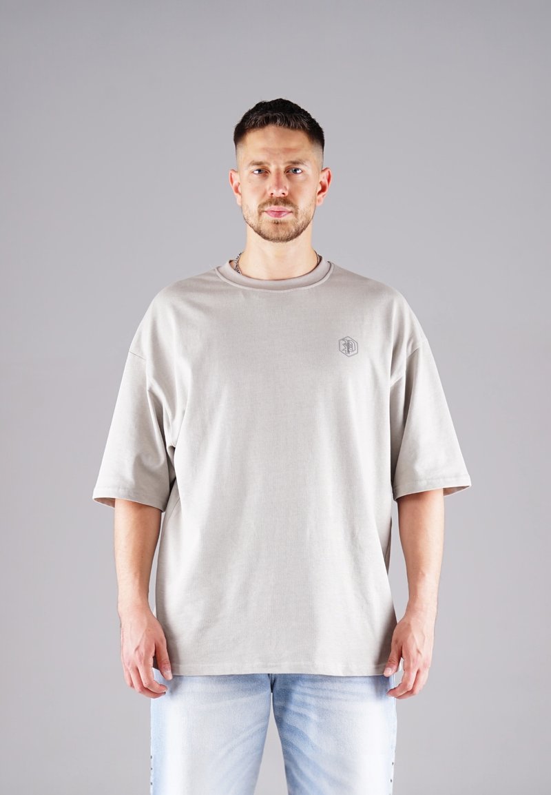 Man with short hair and beard wearing oversized light gray T-shirt and light blue jeans, standing against plain gray background.