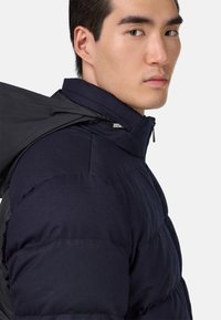 Tumed puffer-jakk, millel on kõrge krae, tekstureeritud viimistlus ja nähtavad õmblused. Lukuga sulgemine ja poleeritud disainielemendid.