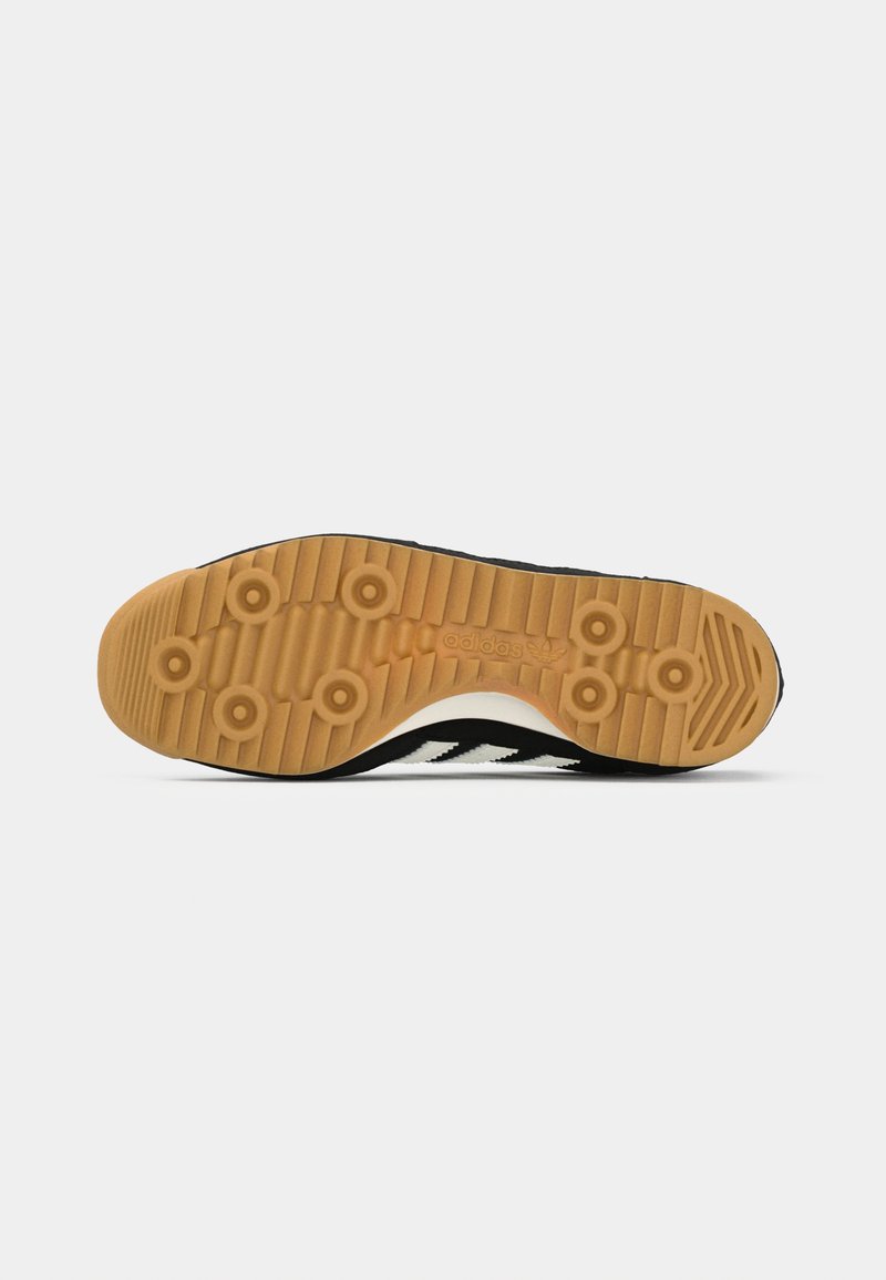 Zapatilla en blanco y negro con suela de goma texturizada, que presenta tres rayas blancas, patrones de pisada redondeados y el logo de Adidas en el lateral.