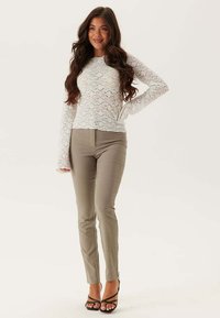 Top lungo a maniche lunghe in pizzo bianco abbinato a pantaloni fitted beige e tacchi neri con cinturino. Dettagli di texture e sovrapposizione visibili nel design del top.