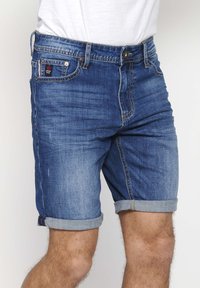 Pantalones cortos de mezclilla en azul medio, con dobladillo enrollado, diseño tradicional de cinco bolsillos y costuras en contraste. Textura suave, estilo casual.