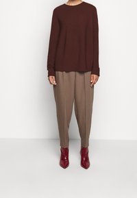 Eine Person trägt einen dunkelbraunen Pullover, lockere beige Hosen und spitze rote Stiefeletten und steht vor einem schlichten weißen Hintergrund.