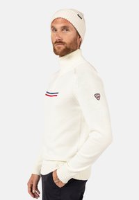 Maglione bianco lavorato a maglia con collo alto, caratterizzato da strisce orizzontali rosse e blu sul petto e un patch con logo sulla manica.