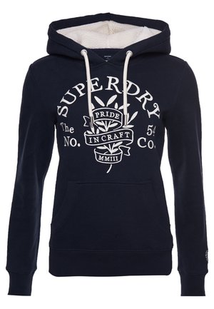 Sweat à capuche bleu marine avec cordons blancs, poche avant, texte blanc « Superdry » et motif végétal avec bannière « Pride in Craft » sur la poitrine.