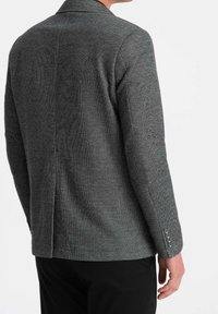 Szary tkany blezer z wyraźną strukturą, patką na klapie, długimi rękawami i mankietami zapinanymi na guziki. Widoczny szew z tyłu.
