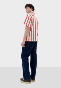 Randig kortärmad skjorta med röda och vita vertikala ränder, kombinerad med mörka denimbyxor och bruna skor på en modell.