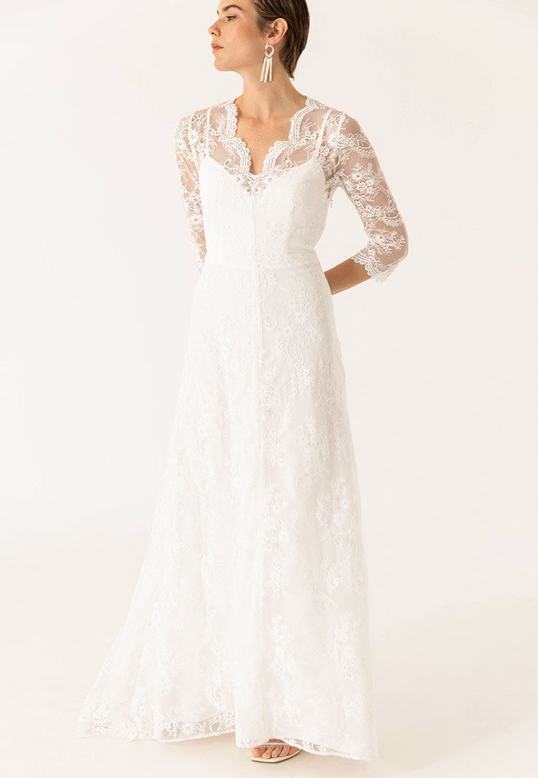 Rochie de mireasă din dantelă albă, cu mâneci lungi din material transparent, modele florale intricate, un corset ajustat și o fustă fluidă cu o ușoară traiectorie.