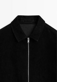 Schwarze Wildlederjacke mit einem eleganten Design, das einen spitzen Kragen und einen silbernen Reißverschluss enthält. Die Textur ist weich und matt, mit minimal sichtbaren Nähten.