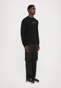 Svart hettegenser med rund hals, med en liten logo-patch; kombinert med svarte cargo-bukser og svarte sportssko.