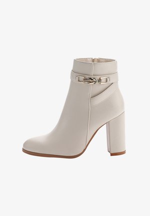 Botín de tobillo de imitación de cuero beige con tacón de bloque, que presenta un acento decorativo de cadena plateada y una textura suave. Diseño de puntera cerrada.