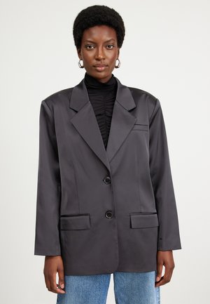 Dunkelgrauer Satin-Blazer mit Reverskragen, zwei Fronttaschen und zwei Knöpfen, kombiniert mit einem schwarzen Oberteil und blauen Jeans.