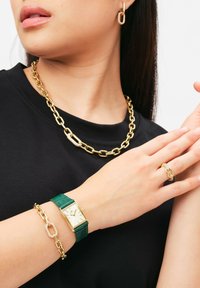 Gold chain necklace, green leather strap watch with gold face, gold bracelet, and ring with chain link detail. Black T-shirt background.