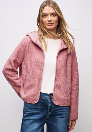 Junge Frau in einer hellrosa strukturierten Zip-Jacke über einem weißen Shirt und blauen Jeans, die vor einem schlichten hellen Hintergrund steht.