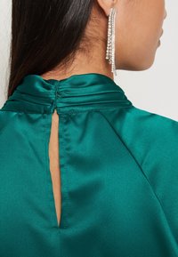 Teal satin top med hög krage och detalj med nyckelhål på baksidan. Modellen bär långa, silvriga örhängen med kaskad av rhinestones.