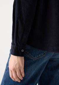 Chemise en velours côtelé bleu marine avec des poignets boutonnés, associée à un jean en denim bleu. Détail de texture visible sur la manche de la chemise.