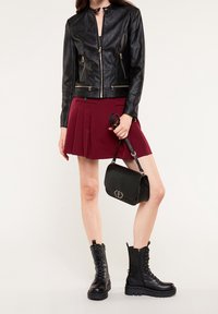 Giacca nera in pelle con tasche con cerniera, gonna plissettata bordeaux, borsa nera con dettaglio metallico e stivaletti da combattimento neri con lacci.