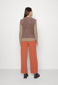 Colete castanho peludo sobre um top bege com riscas, combinado com umas calças largas de cor ferrugem e uns sapatos pretos robustos. Fundo minimalista.