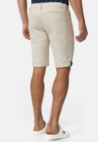Beżowe bawełniane szorty z podwiniętymi nogawkami, dwoma tylnymi kieszeniami i dopasowanym krojem, noszone z białymi, casualowymi sandałami. Gładka tekstura, swobodny styl.