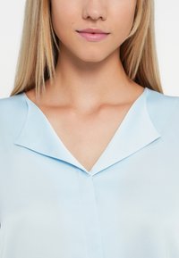 Svijetloplava bluzica s glatkom teksturom, s v-izrezom i suptilnim naborima na izrezu.