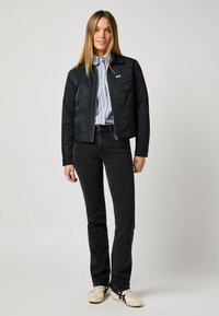 Schwarze gesteppte Jacke mit Reißverschluss, darunter ein blau-weiß gestreiftes Hemd, kombiniert mit schwarzen Jeans und weißen Sneakern mit schwarzen Akzenten.