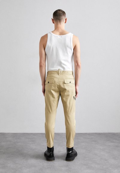 Pantaloni cargo beige con due tasche posteriori, vestibilità dritta e discreto dettaglio del logo di lato, abbinati a una canotta bianca e scarpe nere.