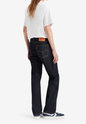 Personne portant un jean bleu foncé droit, un t-shirt blanc court et des baskets bleu et blanc, debout de dos.