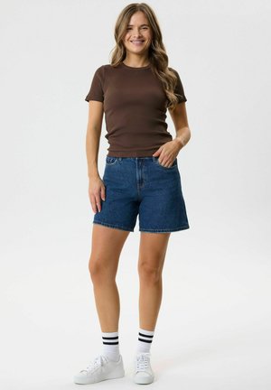 Femme souriante, portant un t-shirt marron à manches courtes, un short en jean bleu, des baskets blanches et des chaussettes blanches à rayures noires, debout devant un fond uni.