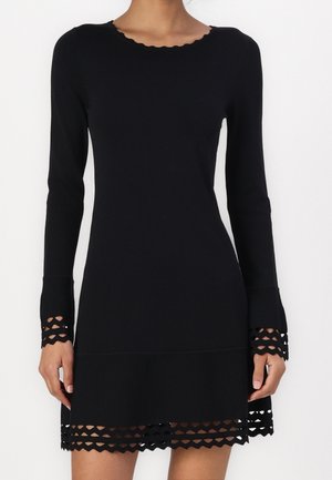 Sort langærmet kjole med bølget halsudskæring og bølgede gitterudskæringsdetaljer på ærmekanter og underkant, båret af en stående person.