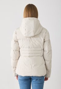 Doudoune beige clair avec capuche, présentant des sections matelassées et une silhouette cintrée. Matériau doux avec une texture lisse et mate.